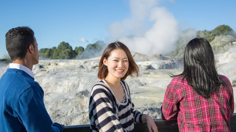 Three students are looking at a geyser in Rotorua. The middle student is facing the camera, smiling.