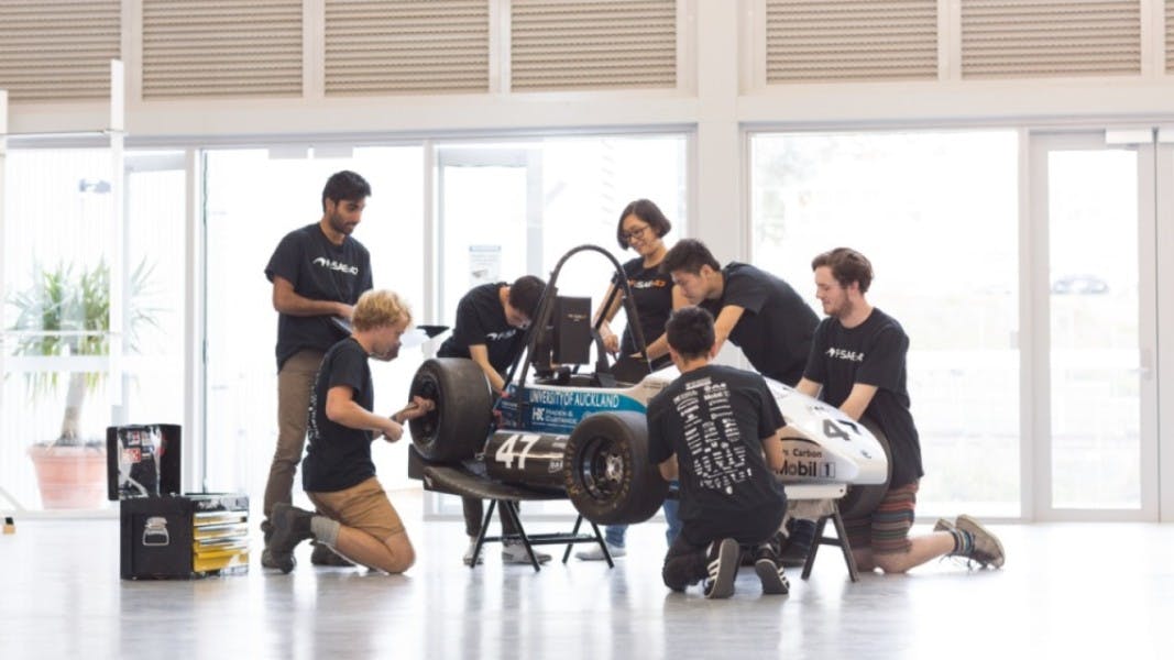 A group of students are gathering around a race car prototype while carrying out engineering work and calculations on it