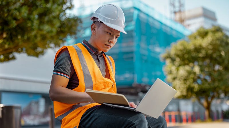 A student in a hard hat and high-vis vest works on a laptop at a construction site. 