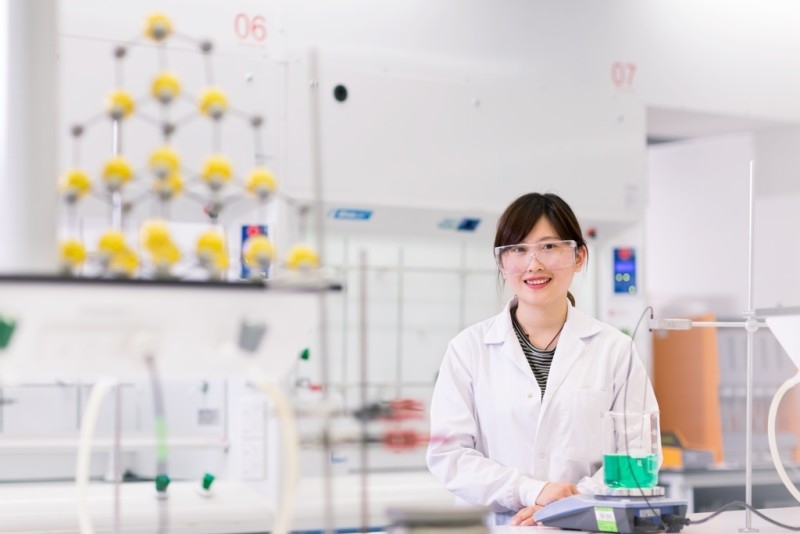 A student in lab coat and safety glasses stands in a university science lab.