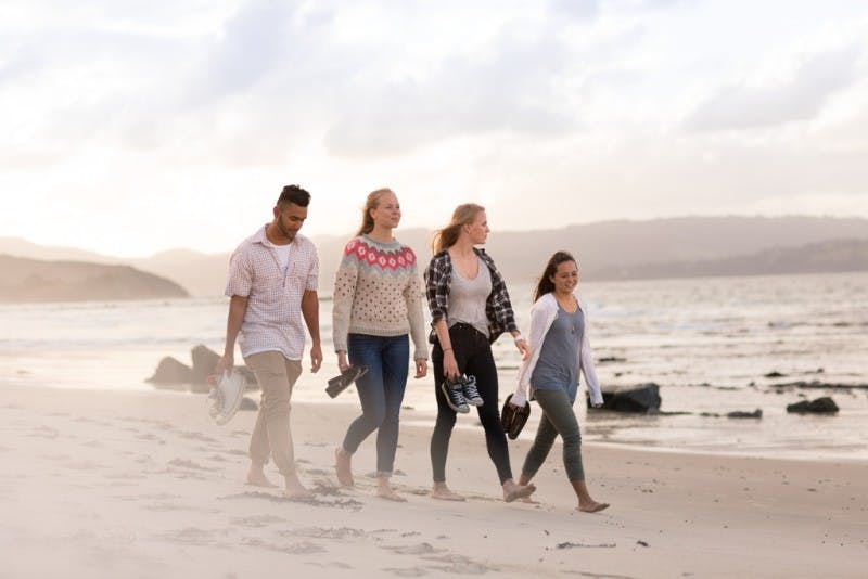 Four international students are walking down a beach in bare feet, holding their shoes and smiling together