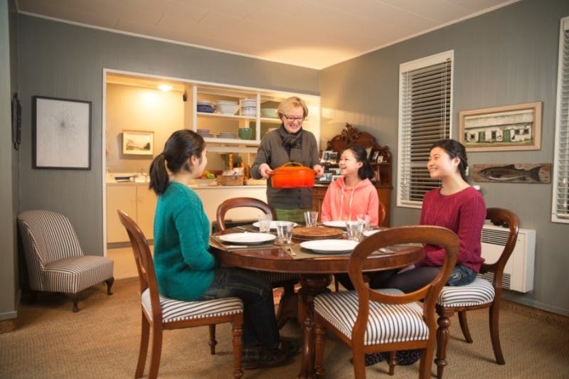 Three international students sitting around a table are being served a meal by their homestay mother from New Zealand