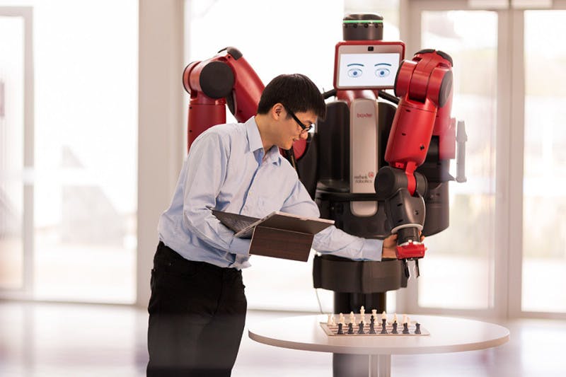 An Information Technology student holding a laptop is working with a robot in front of a chessboard