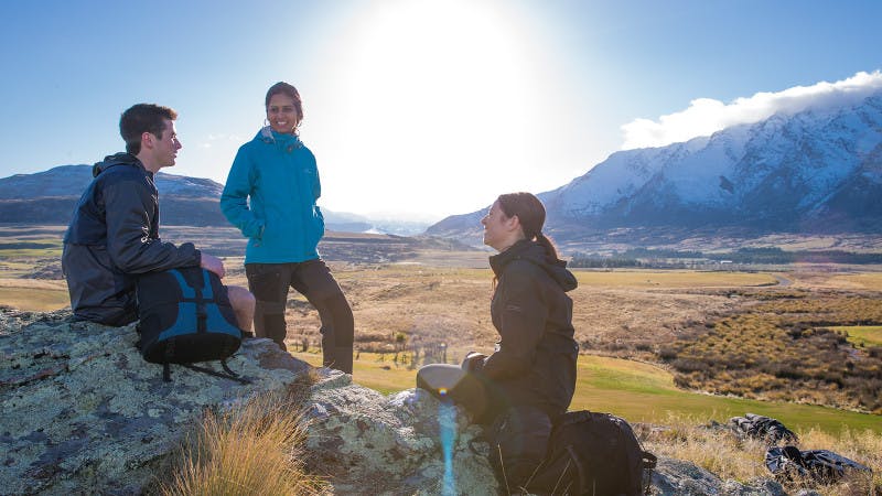 A group of students hiking in Queenstown are taking a break on a rock overlooking a mountain valley.