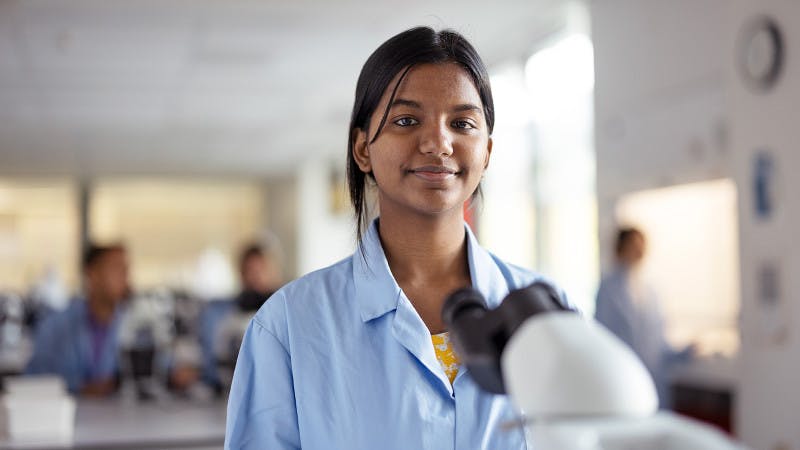 A New Zealand international student in a lab coat, standing behind a microscope