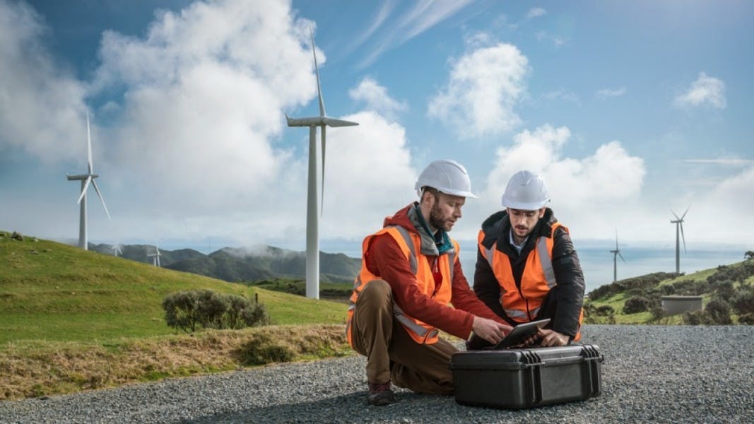 Two engineering students in full safety and high-vis gear look at a tablet screen while near wind turbines on a hill